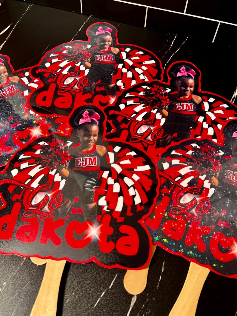 May include: A collection of red, black, and white cheerleading-themed cutouts. Each cutout features a young girl in a cheerleading uniform with the text "Go Dakota". The cutouts have wooden sticks attached.