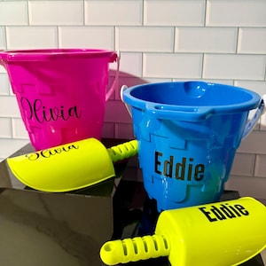May include: Two personalized beach buckets, one pink and one blue, with matching yellow shovels. The pink bucket has "Olivia" in black script, and the blue bucket has "Eddie". The shovels are also personalized with the names.