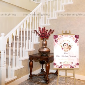 Pode incluir: Bem-vindo &agrave; Cerim&ocirc;nia de Alimenta&ccedil;&atilde;o de Arroz da Clara, um cartaz com um design floral rosa e bord&ocirc;. O cartaz apresenta uma foto de uma menina pequena com um vestido branco e uma faixa de cabe&ccedil;a rosa. O texto do cartaz diz "Bem-vindo &agrave; Cerim&ocirc;nia de Alimenta&ccedil;&atilde;o de Arroz da Clara. Estou pronta para minha primeira tigela de arroz."