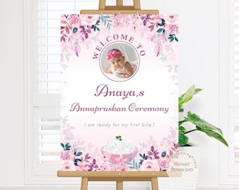 May include: A welcome sign for a baby's Annaprashan Ceremony. The sign features a photo of a baby, floral accents, and the text "Welcome to Anaya's Annaprashan Ceremony. I am ready for my first bite!" in shades of pink and purple.