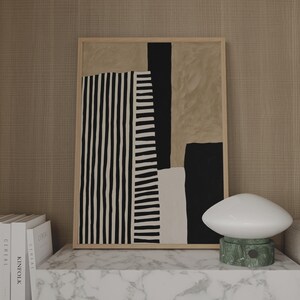 May include: A framed rectangular wall art piece featuring an abstract composition of vertical black and white stripes, solid black blocks, and beige shapes. The artwork is displayed in a light wood frame, positioned next to a white mushroom-shaped lamp.