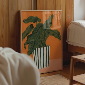 May include: A framed rectangular wall art print featuring a dark green leafy plant in a white pot with vertical dark green stripes. The background of the illustration is a solid orange color, and the print is encased in a light-toned wooden frame.