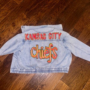 May include: Light blue denim jacket with the words "Kansas City Chiefs" in red and yellow lettering across the back. The jacket has long sleeves and a button-up front.