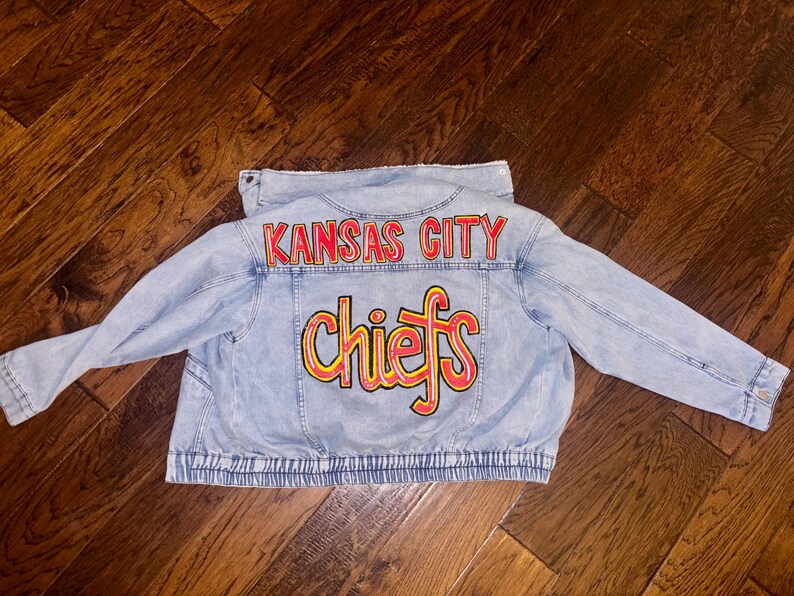 May include: Light blue denim jacket with the words "Kansas City Chiefs" in red and yellow lettering across the back. The jacket has long sleeves and a button-up front.