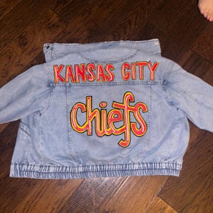 May include: Light blue denim jacket with the words "KANSAS CITY" arched across the upper back in red lettering. Below, the word "chiefs" is written in yellow with a red outline. The jacket has a ribbed hem.
