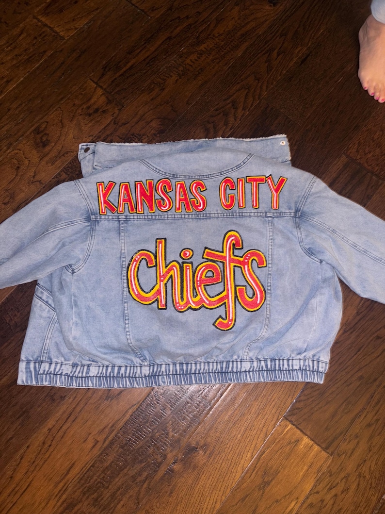 May include: Light blue denim jacket with the words "KANSAS CITY" arched across the upper back in red lettering. Below, the word "chiefs" is written in yellow with a red outline. The jacket has a ribbed hem.