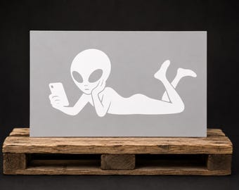 May include: A rectangular grey sign featuring a white graphic of an alien lying on its stomach, propped up on one elbow while looking at a smartphone. The alien has a large oval head, dark almond-shaped eyes, and bent legs.
