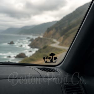 May include: Black mushroom decal on a car dashboard, with a scenic coastal road and ocean view in the background. The decal is a silhouette of several mushrooms. The text "Boston Print Co." is visible on the dashboard.