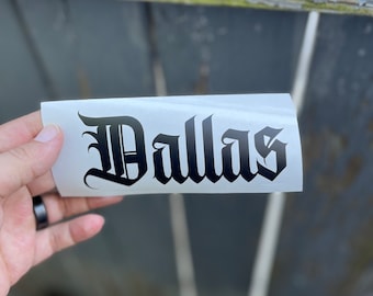 May include: A rectangular white vinyl decal featuring the word Dallas printed in a bold black gothic-style font. The lettering is centered on the adhesive backing, showcasing sharp, ornate edges characteristic of traditional blackletter typography.
