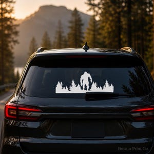 Op de afbeelding: Een witte vinyl sticker van Bigfoot die voor een bosscène staat, aangebracht op de achterruit van een zwarte SUV. De sticker toont een silhouet van het legendarische wezen en groenblijvende bomen.