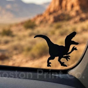May include: A black silhouette decal of a velociraptor dinosaur in a running pose, applied to the surface of a vehicle window. The dinosaur is depicted with its tail extended, claws visible, and mouth slightly open, facing toward the right side.