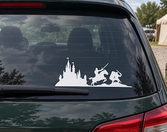May include: A white vinyl decal applied to a car rear window, featuring a silhouette design of a medieval castle, a knight riding a horse, and a soldier on foot holding a sword and shield, all positioned on a rocky landscape base.