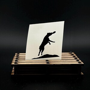 Op de afbeelding: Zwarte silhouet van een hond die springt om een frisbee te vangen. De silhouet is gedrukt op een witte sticker.