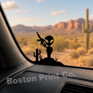 Könnte beinhalten: Eine schwarze Silhouette eines Aliens, das ein Gewehr hält, neben einem Kaktus. Das Alien ist vor einer Wüstenlandschaft mit Bergen und blauem Himmel platziert. Der Text "Boston Print Co." ist sichtbar.