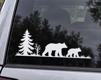 Puede incluir: Un adhesivo de vinilo blanco aplicado en la ventana de un vehículo, con un diseño de silueta de un oso grande caminando detrás de un osezno, acompañado de un pino alto y un árbol más pequeño sobre una línea de suelo herboso.