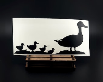 May include: A black silhouette decal featuring a large duck followed by four smaller ducklings standing on a grassy mound. The decal is displayed on a rectangular wooden stand with a slatted design and dark brown finish.