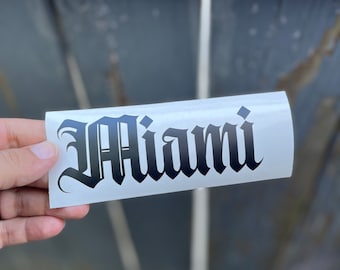 May include: A rectangular white vinyl decal featuring the word Miami printed in a bold, black gothic-style font. The lettering has a sharp, angular design with decorative flourishes on the capital M and lowercase letters.