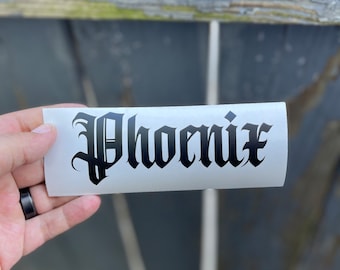 May include: A rectangular white vinyl decal featuring the word Phoenix printed in a bold black gothic-style font. The lettering is centered on the adhesive backing, showcasing sharp, ornate edges characteristic of traditional blackletter typography.