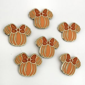 Op de afbeelding: Zes oranje pompoenvormige emaille pins met gouden randen en glinsterende rood-oranje strikken. De pins zijn gerangschikt op een wit oppervlak. Het ontwerp doet denken aan een populair stripfiguur.