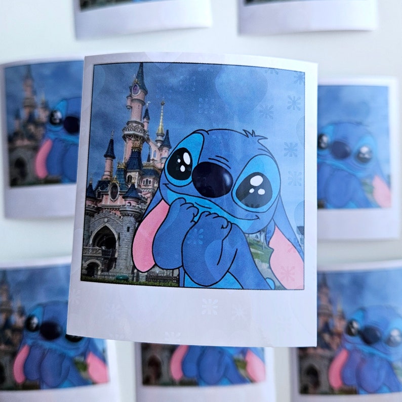 May include: A blue Stitch character with big eyes and a pink nose is looking at the camera. The character is in front of a cartoon image of a castle with pink and white towers. The image is in a white frame with a snowflake pattern.