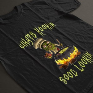 Puede incluir: Camiseta negra con un personaje de chef zombie verde cocinando en una olla sobre un fuego. El texto "WHAT'S COOKIN' GOOD LOOKIN'" está escrito en una fuente gótica.