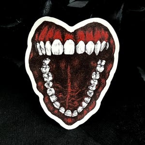 May include: A sticker depicting a heart-shaped open mouth with red interior and white teeth. The sticker has a white border and is set against a black background.