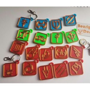 May include: A collection of colorful keychains featuring various symbols. The keychains have a square shape with rounded corners and a small loop for attachment. The symbols are in gold, with a red, green, or blue background. Some keychains have a silver clasp.