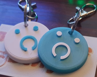 Puede incluir: Dos llaveros con un diseño de cara sonriente blanca y azul. Un llavero es blanco con una cara sonriente azul y el otro es azul con una cara sonriente blanca.