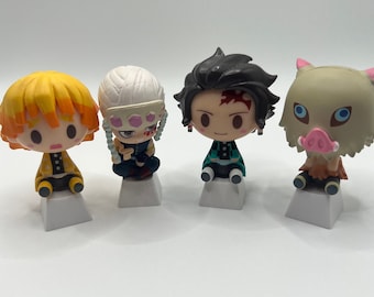 May include: Four small, colorful figurines of characters from the anime series Demon Slayer. The figurines are posed sitting down and are on small white bases. The characters are Tanjiro Kamado, Zenitsu Agatsuma, Inosuke Hashibira, and Nezuko Kamado.