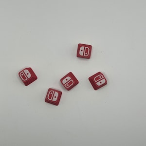 Könnte beinhalten: Fünf rote Keycaps mit einem weißen Nintendo Switch-Logo. Die Keycaps sind in einem Halbkreis auf einer weißen Oberfläche angeordnet.