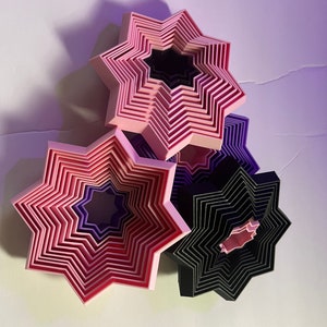 May include: A set of eight star-shaped cookie cutters in various shades of pink, purple, and black. The cutters are nested inside each other, creating a layered effect.