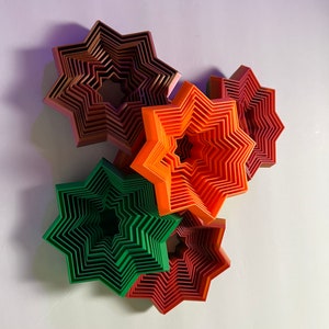 May include: Five layered star-shaped wall decorations in shades of red, orange, and green. The stars are layered on top of each other, creating a three-dimensional effect.