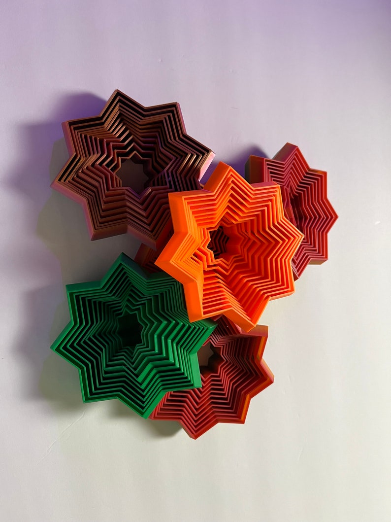 May include: Five layered star-shaped wall decorations in shades of red, orange, and green. The stars are layered on top of each other, creating a three-dimensional effect.