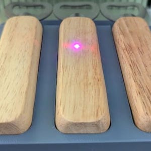 Könnte beinhalten: Drei Flaschenöffner aus Holz mit hellfarbiger Holzmaserung sind in einem blauen Halter angeordnet. Ein kleiner, leuchtend rosa Laserpunkt ist auf dem mittleren Öffner sichtbar. Die Öffner haben eine rechteckige Form mit abgerundeten Kanten.