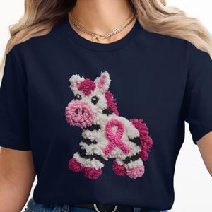 May include: Navy blue t-shirt featuring a whimsical, textured zebra design with a pink ribbon, symbolizing breast cancer awareness. The zebra has black and white stripes, pink mane, and a pink nose. The design is centered on the shirt.