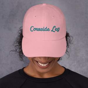 May include: A light pink baseball cap with the embroidered text "Cowside Leg" in teal. The cap has a curved brim and a classic six-panel design. The hat is a solid color and is suitable for casual wear.