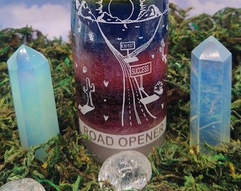 May include: A glass candle with a rainbow design and the words "Abre Camino" and "Road Opener". The candle is surrounded by crystals and moss.