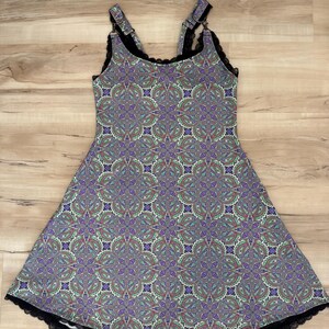 May include: A sleeveless dress featuring a repeating purple, green, and blue paisley-inspired floral pattern. The garment has black lace trim along the neckline and hem, with adjustable shoulder straps connected by metal rings and clips.