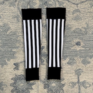 May include: Two black and white striped leg warmers. Each has a solid black band at the top and bottom, with vertical white stripes on a black background. The leg warmers are on a patterned rug.