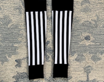 May include: Pair of black and white striped leg warmers. Each leg warmer has a solid black band at the top and bottom, with vertical white stripes on a black background. The leg warmers are laying on a patterned rug.