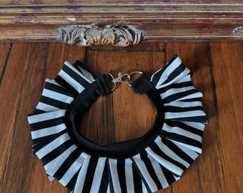 May include: A black and white striped collar with a silver clasp. The collar has a ruffled design and is made of fabric. The item is laying on a wooden surface.