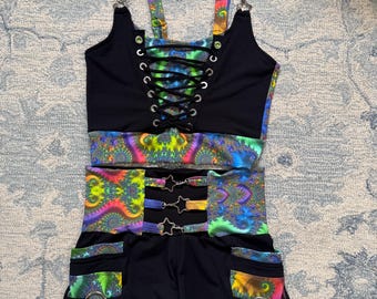 May include: A two-piece outfit featuring a black crop top with a lace-up front and colorful straps. The matching shorts have pockets and a vibrant, psychedelic pattern in shades of blue, green, yellow, and purple. Metal hardware accents the design.