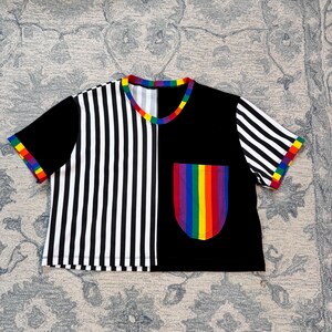 May include: A short-sleeved, cropped t-shirt with a unique design. The shirt is split vertically, with one side featuring black and white stripes and the other solid black. A rainbow-colored pocket and trim add a pop of color.