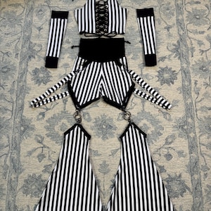 May include: A black and white striped outfit displayed on a patterned rug. The ensemble comprises a halter top with a lace-up front, shorts, arm warmers, and leg pieces. The stripes are vertical and consistently spaced.