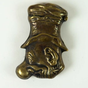 May include: A brass match safe shaped like a man's head wearing a hat. The man's face is detailed with a large nose and a wide mouth. The hat is closed and has a small hinge.