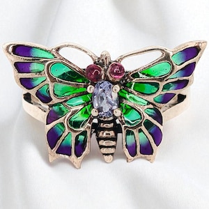Puede incluir: Anillo de mariposa dorado con alas de esmalte verde y morado. El anillo presenta una piedra central ovalada, azul claro y dos pequeñas piedras rosas. El diseño de la mariposa es detallado, sobre un fondo blanco.