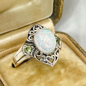 May include: An antique silver ring with a large oval opal center stone and two smaller green stones, set in an ornate filigree design. The ring is displayed in a vintage leather jewelry box with a velvet interior.