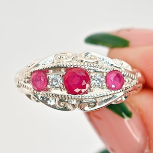May include: A silver ring with three bright red gemstones and smaller clear stones. The ring has an ornate, filigree design along the band. The gemstones are set in a row, creating a classic and eye-catching piece of jewellery. The ring's design evokes a vintage aesthetic.