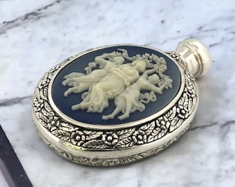 May include: A silver oval-shaped perfume bottle with a detailed floral design around the edge. The bottle features a cameo of three women in white on a blue background.