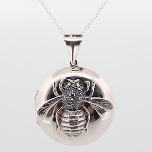 May include: A silver-colored locket necklace with a detailed bee pendant. The bee has textured wings and body, and the locket is round. The necklace chain is delicate and silver.